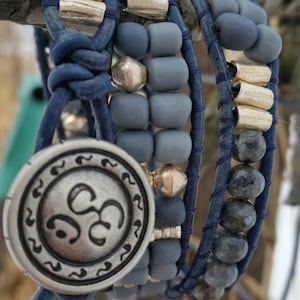 以下が含まれることがあります： 青い革紐、灰色のビーズ、銀色のビーズ、そしてオームのシンボルが刻まれた銀のチャームが付いた、マルチストランドブレスレット。