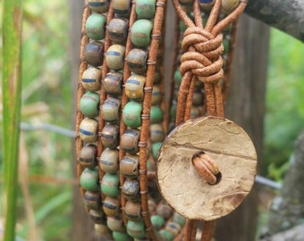 May include: Wrap bracelet with green, brown, and blue beads. The bracelet is made of brown leather and fastens with a wooden button. The beads are strung on the leather, creating a layered effect. The bracelet is wrapped around a branch.