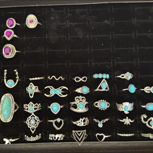 May include: A collection of turquoise and silver rings displayed in a black velvet-lined box. The rings feature various designs, including floral, geometric, and animal motifs.