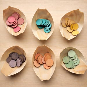 Peut inclure: Six plateaux en bois en forme de feuille, chacun rempli de petits disques ronds en bois. Les disques sont de différentes couleurs, notamment rose, sarcelle, jaune, marron, orange et vert. Les plateaux sont en bois clair.