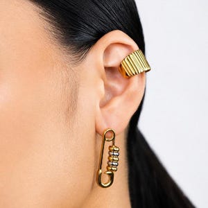 Könnte beinhalten: Goldfarbene Ohrklemme und Sicherheitsnadel-Ohrringe. Die Ohrklemme hat ein geripptes Design, während die Ohrringe eine Sicherheitsnadelform mit einem Perlenakzent aufweisen. Der Schmuck wird an einem Ohr getragen, mit schwarzem Haar sichtbar.