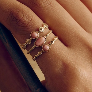 May include: Three gold rings with pink oval gemstones are displayed on a finger. The rings have intricate designs, with one featuring a row of small beads. The gemstones are a soft, pastel pink color. The rings are set against a warm, neutral background.
