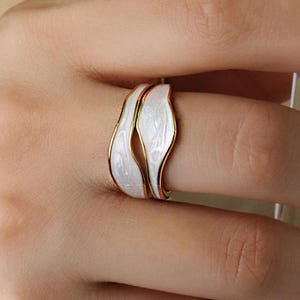 Puede incluir: Primer plano de un anillo blanco y dorado con un diseño único y fluido. El anillo presenta una incrustación de esmalte blanco con bordes dorados. El anillo está en un dedo.