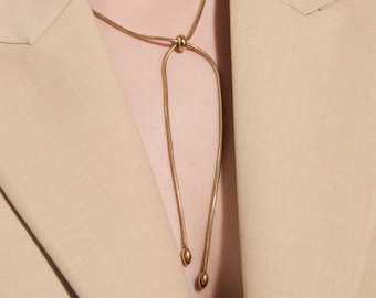Può includere: Una collana color oro dal design unico. La collana presenta un elemento annodato nella parte superiore, con due fili pendenti che terminano con piccoli pendenti arrotondati. La collana è indossata sopra un blazer beige.