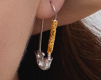 Könnte beinhalten: Ein silberner und goldener Ohrring in Form einer Sicherheitsnadel. Der goldene Teil des Ohrrings hat ein verziertes, geschnitztes Design. Der Ohrring befindet sich im Ohr einer Person mit braunem Haar.
