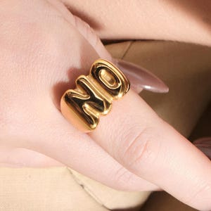 Pode incluir: Um anel dourado com a palavra "NO" escrita em uma fonte arrojada e arredondada. O anel é usado em um dedo, com a mão e parte do braço visíveis. O anel tem uma superfície brilhante e reflexiva.