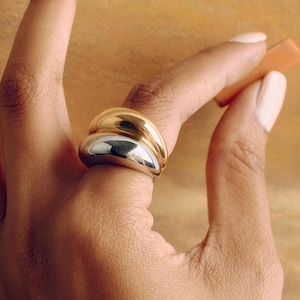May include: A two-toned ring featuring a smooth, rounded gold band stacked on top of a smooth, rounded silver band. The ring has a polished, reflective finish and a thick, chunky profile that sits prominently on the finger.