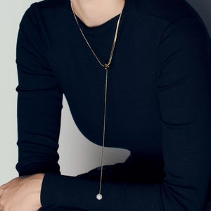 May include: A gold-colored lariat necklace with a single pearl pendant. The necklace is draped over a black long-sleeved top. The design features a delicate chain and a simple, elegant style.