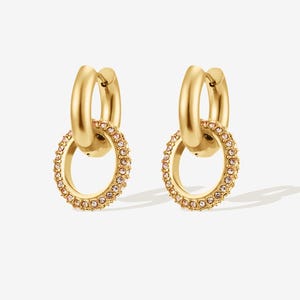 May include: A pair of gold-colored hoop earrings. Each earring features a smaller, dangling hoop embellished with small, pink-toned crystals. The earrings have a polished, shiny finish and are set against a white background.