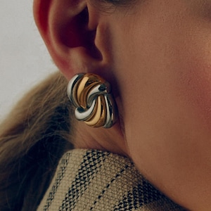 May include: A close-up of a gold and silver-toned earring. The earring is a clip-on style, featuring a swirling design with alternating gold and silver metal segments. The earring is worn on a person's ear.