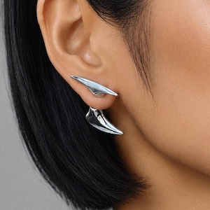 May include: A silver-toned ear cuff featuring two distinct, pointed, curved elements. The upper piece rests along the outer edge of the ear cartilage, while the lower piece extends downwards, creating a layered, sculptural appearance with a polished finish.