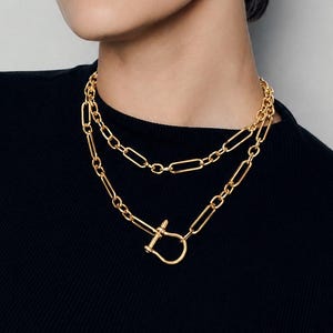 May include: A gold-toned double-layered chain necklace featuring elongated oval links. The lower chain includes a prominent U-shaped shackle clasp closure with a horizontal bar pin, creating a distinct focal point against the black fabric.