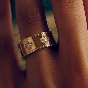 May include: A gold ring featuring a repeating clover pattern and small, clear gemstones. The ring has a textured, scalloped edge. The background is a blurred landscape. The ring is worn on a finger.