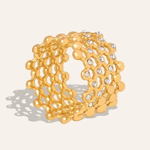Puede incluir: Un anillo dorado con diseño de panal. El anillo presenta pequeñas piedras preciosas transparentes engastadas en el patrón de panal. El anillo se muestra sobre un fondo neutro, proyectando una sombra.