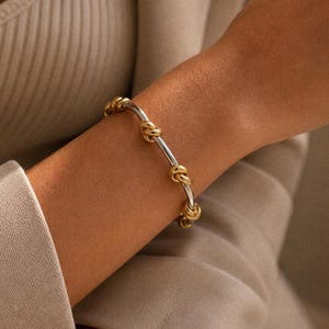 Könnte beinhalten: Ein silbernes und goldenes Armband mit Knotenmuster. Das Armband hat ein silbernes Band mit goldfarbenen Knoten, die gleichmäßig über den Umfang verteilt sind. Das Armband wird am Handgelenk einer Person getragen, mit einem neutralen Hintergrund.