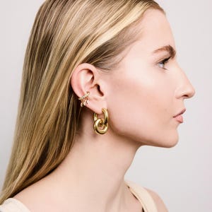 May include: A side profile view of a person wearing gold-colored jewelry. The jewelry includes a pair of large hoop earrings and a decorative ear cuff with small, sparkling embellishments. The person has long, blonde hair.