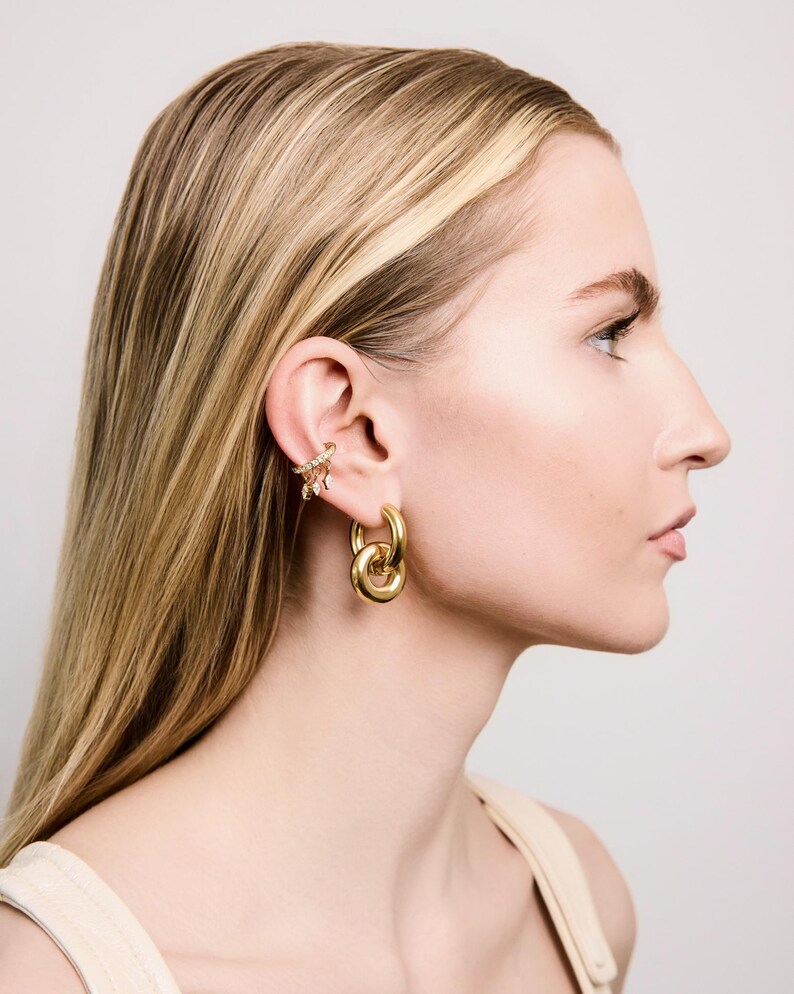 May include: A side profile view of a person wearing gold-colored jewelry. The jewelry includes a pair of large hoop earrings and a decorative ear cuff with small, sparkling embellishments. The person has long, blonde hair.
