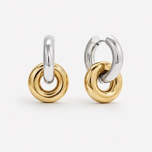 May include: A pair of two-tone hoop earrings. The top portion of each earring is silver, while the lower, circular portion is gold. The earrings are connected by a small hinge. The background is plain white.