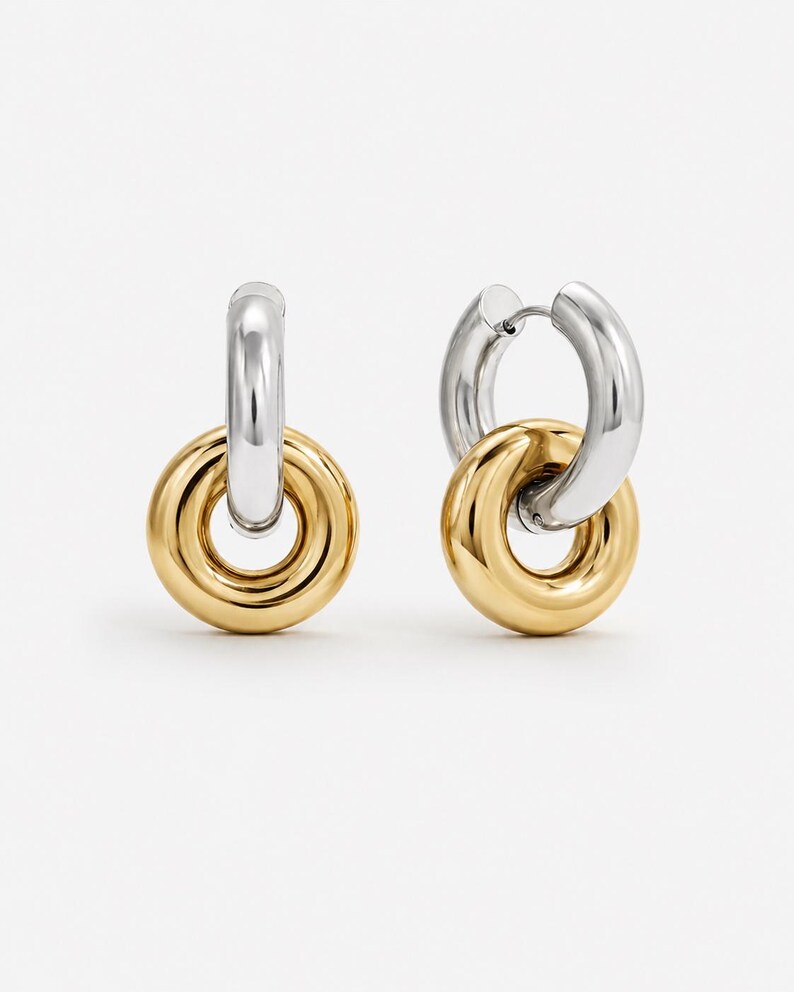 May include: A pair of two-tone hoop earrings. The top portion of each earring is silver, while the lower, circular portion is gold. The earrings are connected by a small hinge. The background is plain white.
