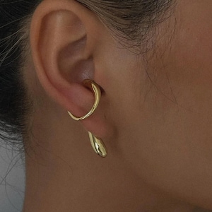 May include: A gold-colored ear cuff and earring set. The ear cuff curves around the upper ear, while the earring dangles below. The jewelry has a smooth, polished finish, reflecting light. The design is modern and minimalist, suitable for everyday wear or special occasions.