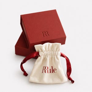 Op de afbeelding: Een rode geschenkdoos met de in reli&euml;f aangebrachte letter "R" en een cr&egrave;mekleurig zakje met trekkoord met het woord "Rule" in het rood gedrukt. De tas heeft rode satijnen linten.