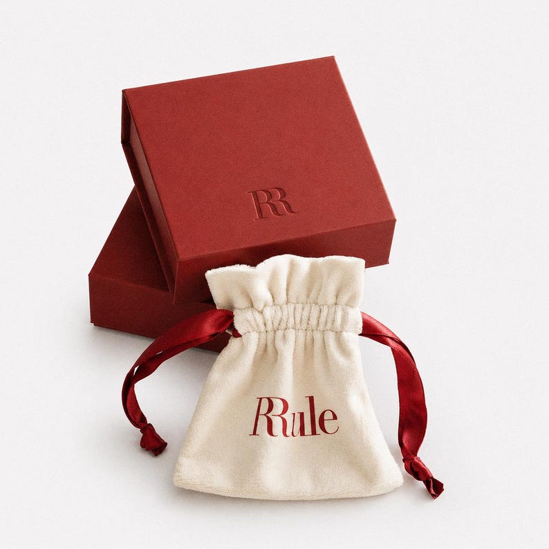 Op de afbeelding: Een rode geschenkdoos met de in reli&euml;f aangebrachte letter "R" en een cr&egrave;mekleurig zakje met trekkoord met het woord "Rule" in het rood gedrukt. De tas heeft rode satijnen linten.