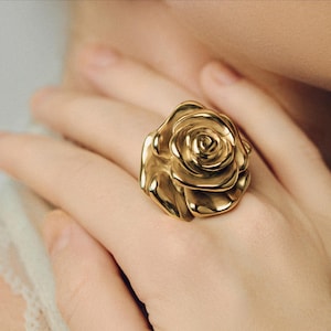 Pode incluir: Um anel dourado em forma de rosa. O anel é usado em um dedo e é o ponto focal da imagem. As pétalas da rosa são detalhadas e texturizadas, criando uma aparência realista. O anel é uma peça de destaque.