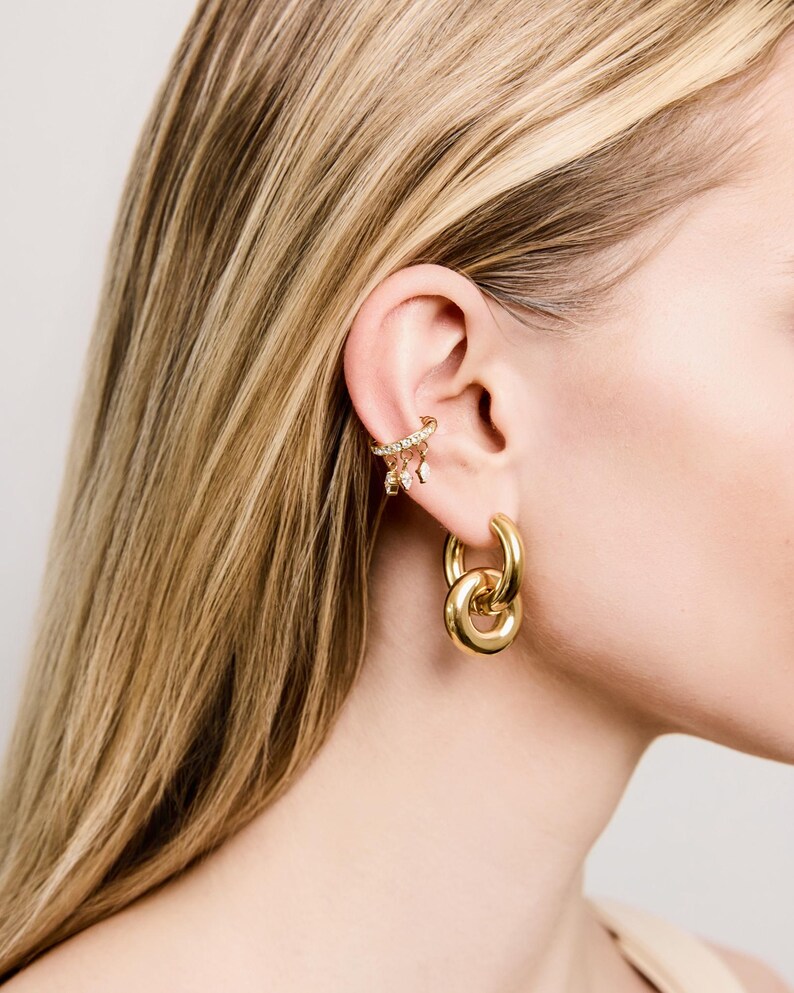 May include: Gold-toned jewelry set featuring a pair of large hoop earrings and a delicate ear cuff. The earrings have a double-loop design, while the ear cuff is adorned with small, sparkling embellishments. The jewelry is displayed on a person's ear.