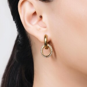 May include: Gold-tone hoop earrings with a smaller, oval-shaped charm. The charm is encrusted with small, clear crystals. The earrings are shown on a person's ear, with their dark hair visible.