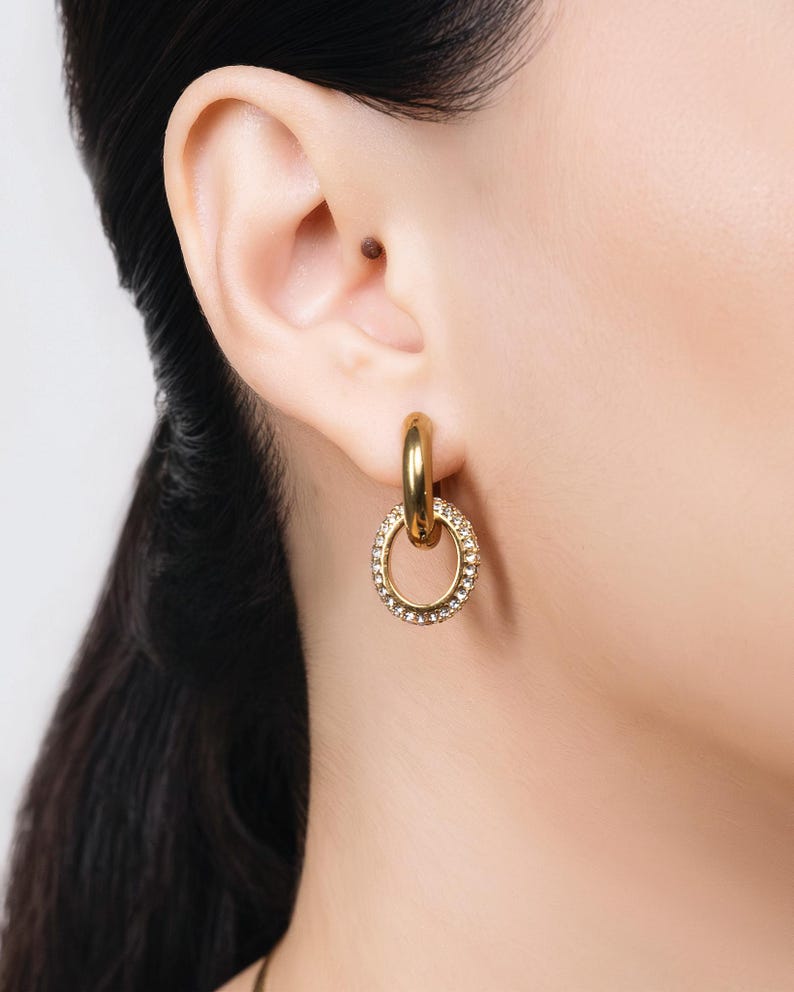May include: Gold-tone hoop earrings with a smaller, oval-shaped charm. The charm is encrusted with small, clear crystals. The earrings are shown on a person's ear, with their dark hair visible.