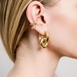 May include: A close-up of a person's ear adorned with gold jewelry. The ear features a delicate ear cuff with small dangling charms and a large, linked hoop earring. The jewelry has a polished, reflective surface.