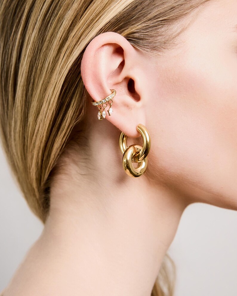 May include: A close-up of a person's ear adorned with gold jewelry. The ear features a delicate ear cuff with small dangling charms and a large, linked hoop earring. The jewelry has a polished, reflective surface.