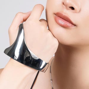May include: A polished silver cuff bracelet with a flared, sculptural design. The bracelet is worn on a wrist, reflecting light and creating a modern, minimalist aesthetic. The jewelry is a statement piece.