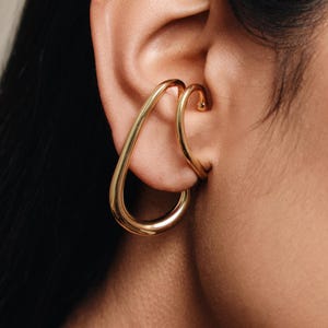Könnte beinhalten: Ein goldfarbener Ohrschmuck mit Tropfendesign. Der Ohrschmuck umschließt das Ohr und erzeugt einen modernen, minimalistischen Look. Der Schmuck besteht aus glänzendem, reflektierendem Metall.
