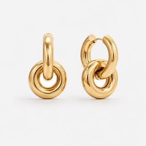 May include: A pair of gold-colored hoop earrings. Each earring features a large, rounded hoop connected to a smaller, circular element. The earrings have a polished, reflective surface, and are set against a white background.