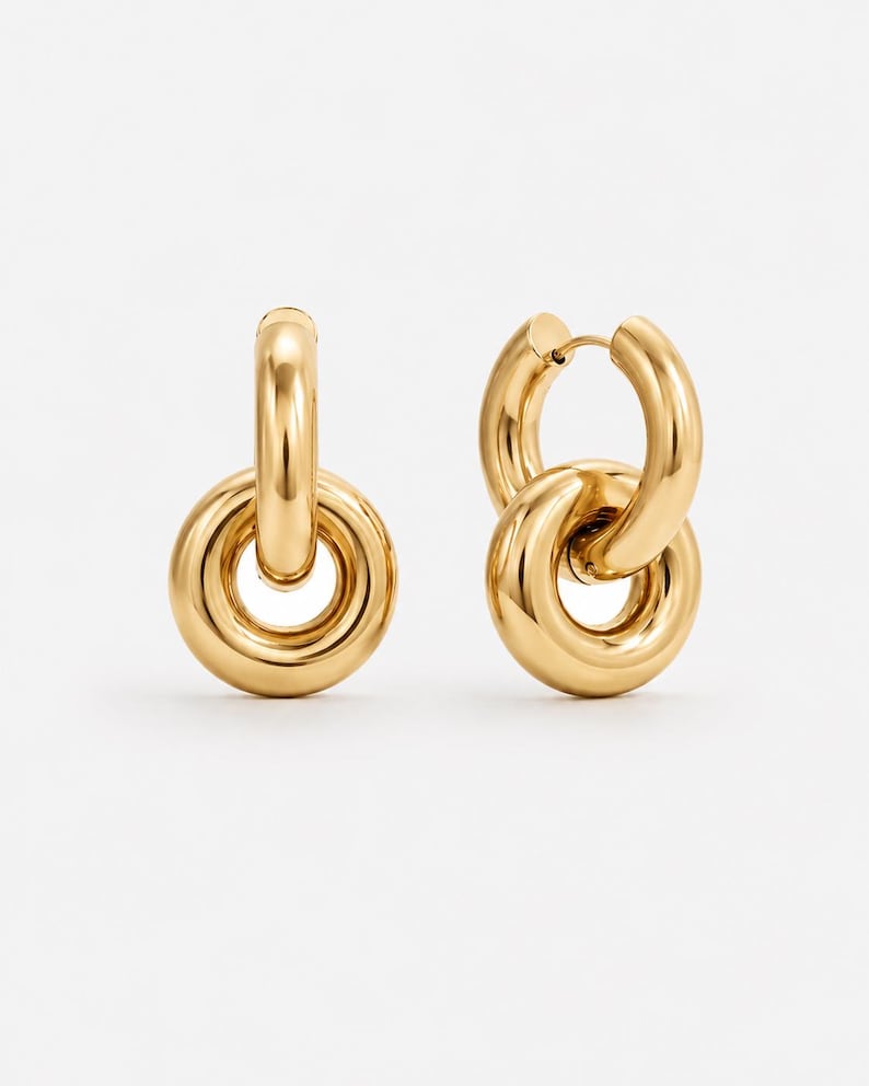 May include: A pair of gold-colored hoop earrings. Each earring features a large, rounded hoop connected to a smaller, circular element. The earrings have a polished, reflective surface, and are set against a white background.