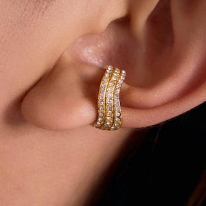 May include: A gold-colored ear cuff with three rows of small, clear crystals. The ear cuff is curved to fit the ear and is worn on the ear cartilage. The jewelry is shown against a blurred background.