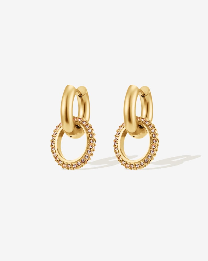 May include: A pair of gold-colored hoop earrings. Each earring features a smaller, dangling hoop embellished with small, pink-toned crystals. The earrings have a polished, shiny finish and are set against a white background.