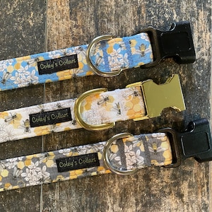 May include: Three dog collars with a bee and flower pattern. The collars are white with yellow and black bees and light blue flowers. The collars have gold and black buckles and the text "Coley's Collars" is printed on each collar.