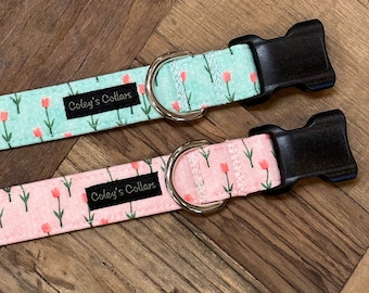 May include: Two dog collars with a floral pattern. The collars are turquoise and pink with a white background and feature small pink tulips. The collars have black buckles and silver D-rings. The collars have a black label that reads "Coley's Collars".