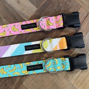 May include: Three dog collars with black plastic buckles and silver metal D-rings. The collars feature colorful patterns: pink with bananas, striped pastel colors, and turquoise with bananas. Each collar has a label that reads "Coley's Collars."