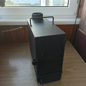 May include: A rectangular black metal portable stove featuring a flat top surface with a circular chimney vent, a sturdy carrying handle, and a front-loading door secured by a curved metal latch mechanism. The stove has a matte finish.