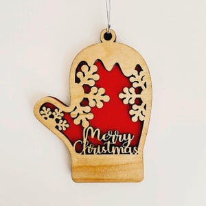 Puede incluir: Adorno navideño de madera con forma de mitón. El mitón es de madera marrón claro con interior rojo. Se ven recortes de copos de nieve y la inscripción "Merry Christmas".