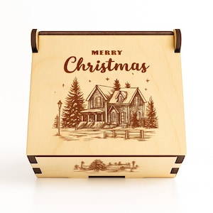 Puede incluir: Una caja de madera con tapa con bisagras, con un diseño grabado con láser. El diseño incluye una casa, árboles y las palabras "MERRY Christmas". La caja es de madera clara con detalles de madera más oscura en las esquinas y bisagras.