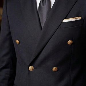 Könnte beinhalten: Ein dunkelblauer, zweireihiger Anzug mit goldenen Knöpfen. Der Anzug hat ein weißes Hemd, eine schwarze Krawatte und ein Einstecktuch mit goldenem Rand. Ein klassischer Anzug für formelle Anlässe.
