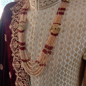 May include: Cream-coloured embroidered formal wear with a multi-strand pearl necklace featuring red accents and gold embellishments. A maroon velvet shawl with gold embroidery is draped over the shoulder. The outfit is displayed on a mannequin.