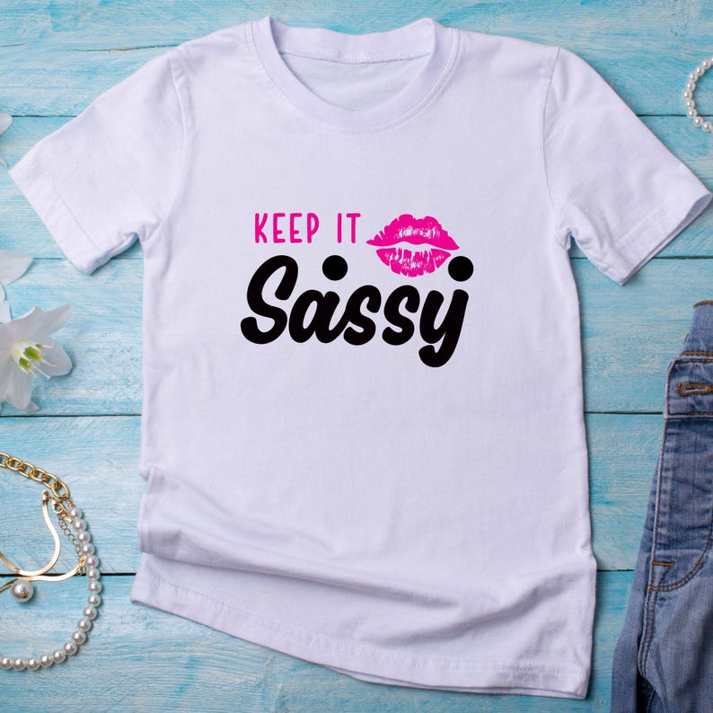 May include: White t-shirt with the text "KEEP IT Sassy" in black and pink. The design includes a pair of pink lips. The shirt is made of a soft material and has short sleeves.