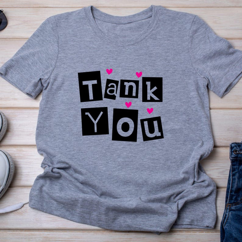 May include: A gray t-shirt with the words "Tank You" in black block letters, with pink heart accents. The shirt is made of a soft material and is suitable for casual wear.