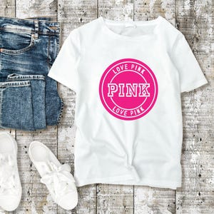 May include: White t-shirt with a pink circular graphic that says "PINK" and "LOVE PINK". The shirt is next to a pair of blue jeans and white sneakers with white laces. The items are on a light wood-grain surface.