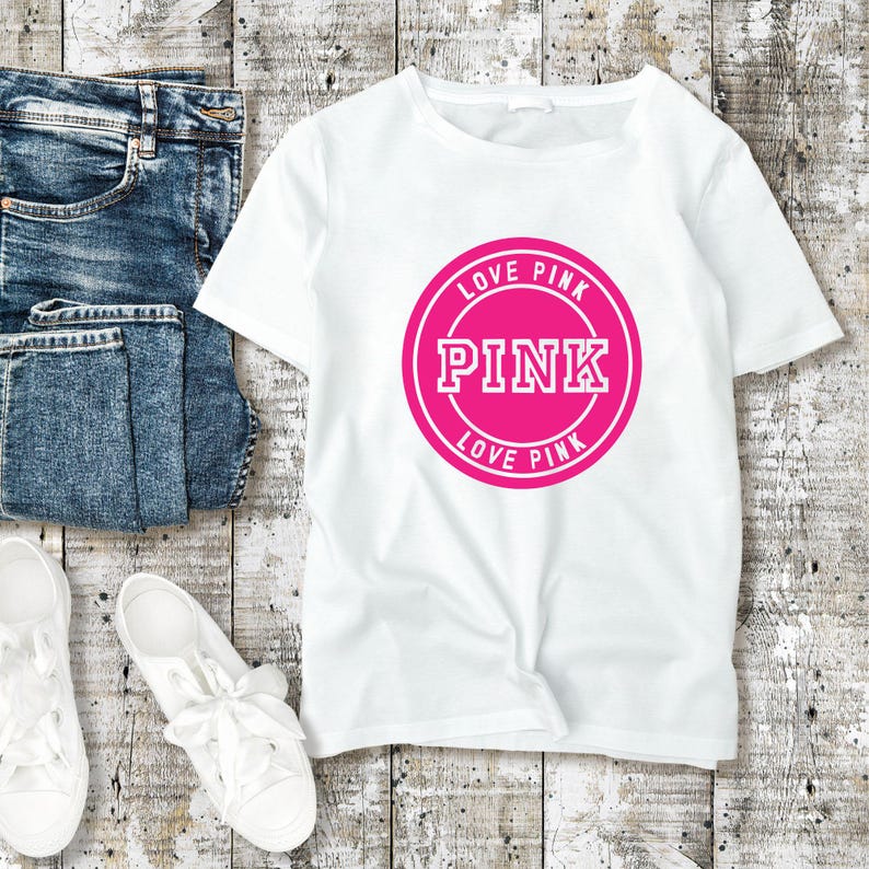 May include: White t-shirt with a pink circular graphic that says "PINK" and "LOVE PINK". The shirt is next to a pair of blue jeans and white sneakers with white laces. The items are on a light wood-grain surface.